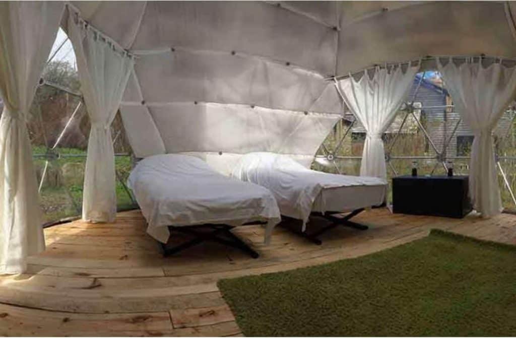 An elegantly furnished bedroom inside a dome tent, featuring a cosy bed and a large, clear skylight for viewing the night sky from the comfort of the room.