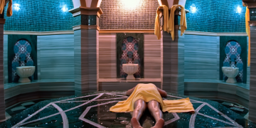 A traditional Turkish Hammam or bathhouse, featuring a central heated stone platform surrounded by marble walls, with a large dome above and several bathing basins.