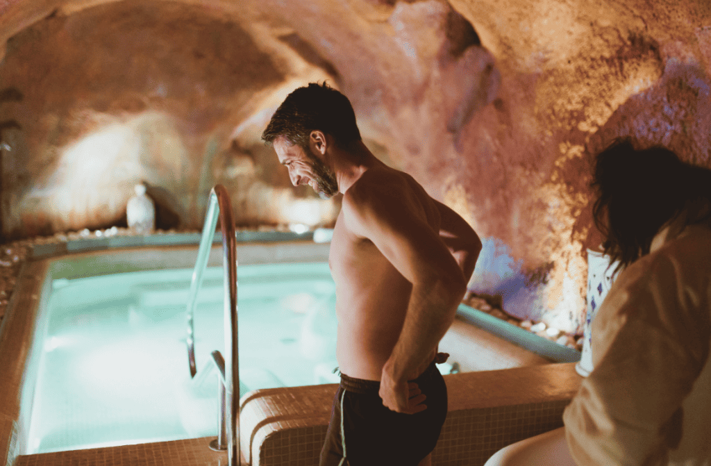 Image of a man stepping into a serene, indoor pool located within a cave-like facility, illustrating a tranquil and unique spa experience with natural stone walls and ambient lighting that creates a peaceful, secluded atmosphere.