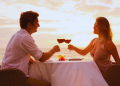 A couple enjoying wine by the beach at sunset, glasses raised in a toast.