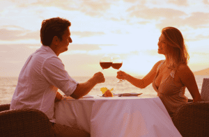 A couple enjoying wine by the beach at sunset, glasses raised in a toast.