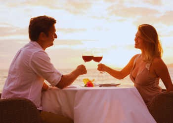 A couple enjoying wine by the beach at sunset, glasses raised in a toast.