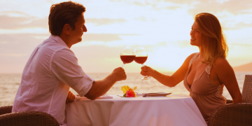 A couple enjoying wine by the beach at sunset, glasses raised in a toast.
