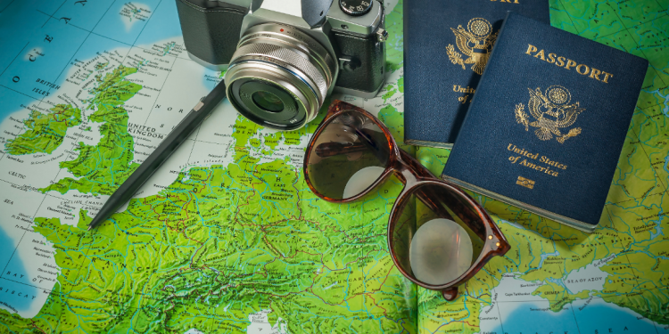 Two American passports with a European map in the background, alongside dark sunglasses and a camera, symbolizing travel preparation.